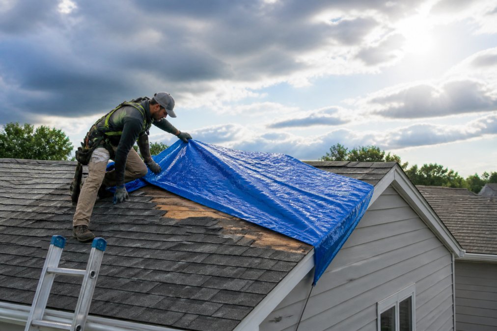 A roofing professional in safety gear securing a blue tarp over a damaged shingle roof section on a suburban home, storm clouds clearing in the background, ladder visible at the roof edge