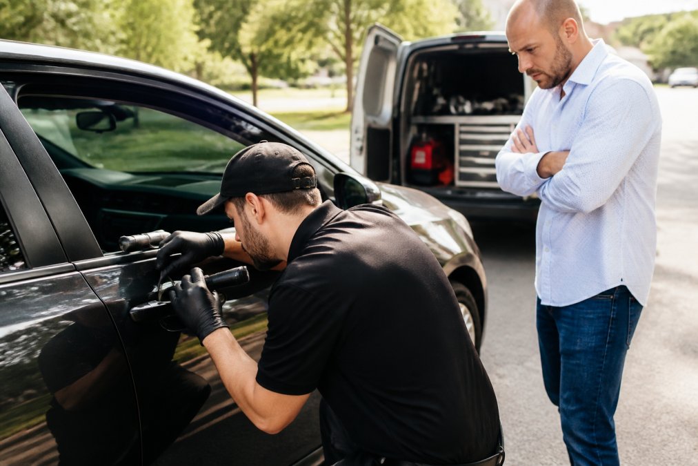 Professional automotive locksmith in uniform using specialized tools to open a vehicle door carefully, demonstrating damage-free entry technique in daylight, with customer standing nearby observing
