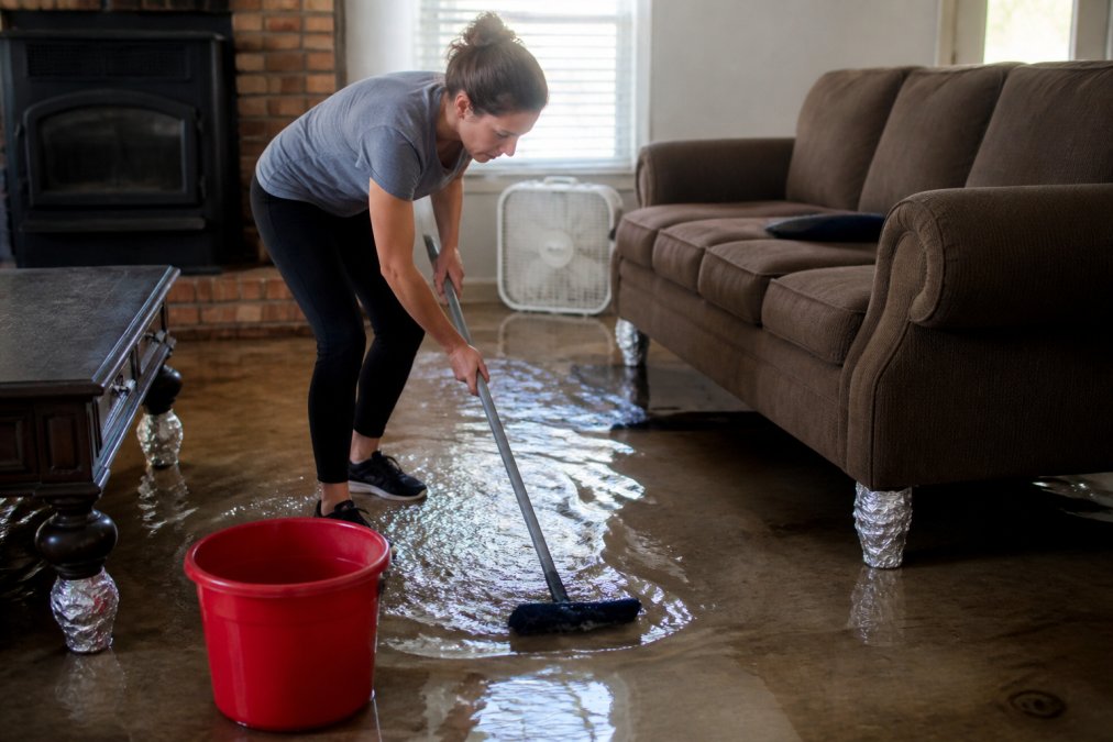 A homeowner in casual clothes using a mop and bucket to soak up standing water in a flooded living room, with furniture legs wrapped in aluminum foil and a box fan running in the background