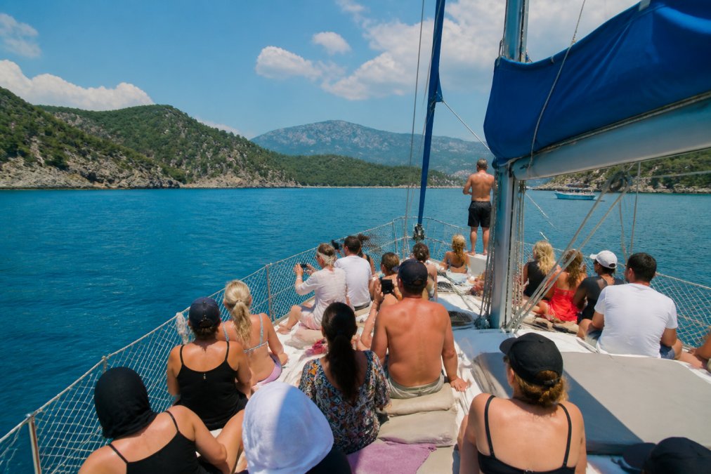 Turkuaz sularında yelkenli tekne, turist grubu güneşte oturmuş, denizde yüzüyor, berrak su ve Fethiye kıyısı arka planda