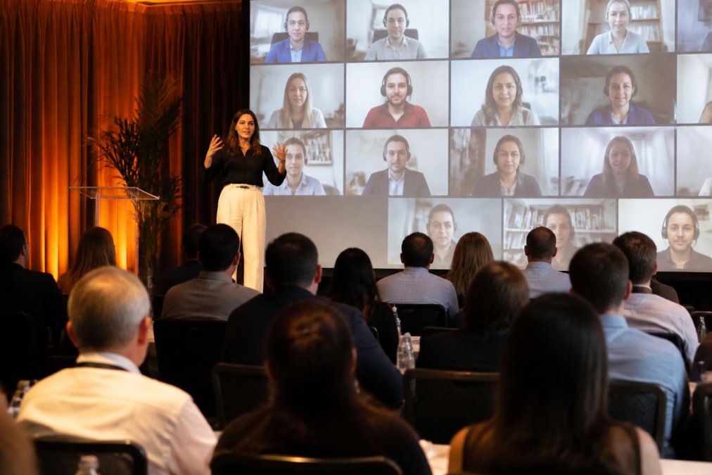 A presenter speaking on stage at a hybrid corporate event, with a large screen behind them showing remote attendees on a video call grid, audience members seated in the foreground under warm event lighting
