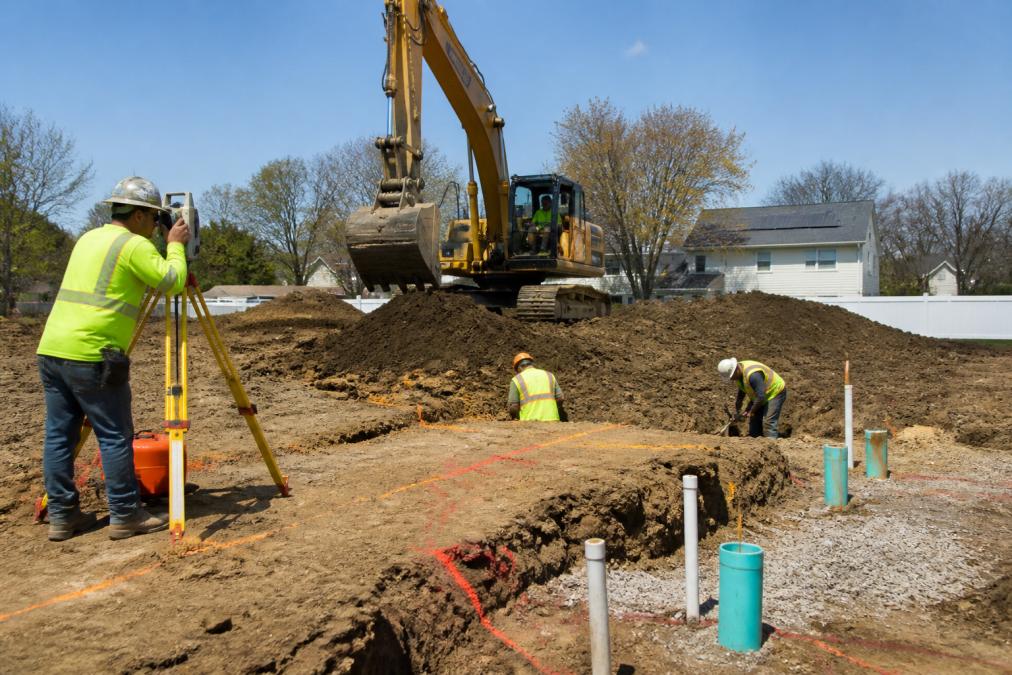 Professional construction crew operating heavy excavation equipment on an active site preparation project, with surveying equipment and marked utility lines visible in the ground, in natural daylight