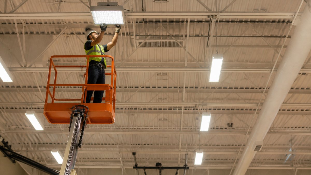 Electrician using a lift to install LED fixtures for energy efficiency upgrades for schools in a gymnasium.
