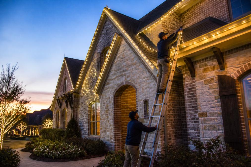 A wide shot of a large, elegant suburban home at dusk, covered in warm white LED holiday lights, with a professional installer on a ladder placing the final string along the roof line