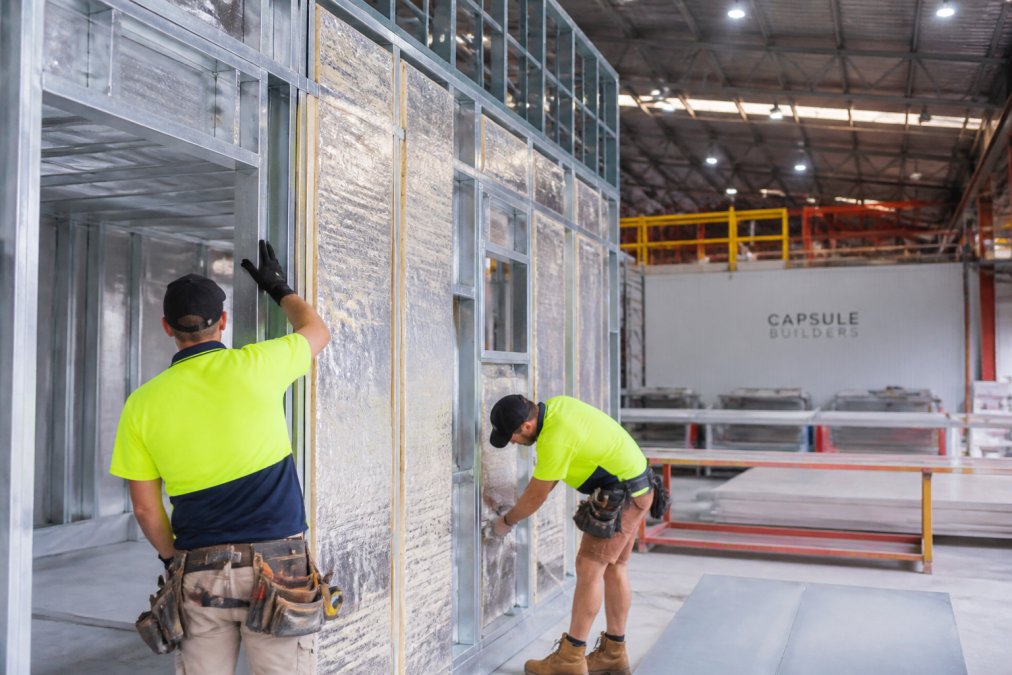 Close-up of a steel-framed modular home under construction in a bright factory, workers in safety gear and hard hats assembling insulated wall panels under overhead lighting