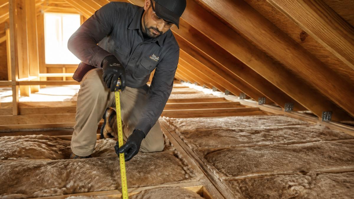 A roofing contractor in work clothes and safety gear measuring attic insulation depth with a tape measure inside a sunlit residential attic, wooden rafters visible, insulation batts laid between joists