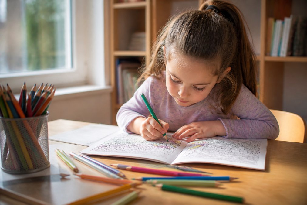 A 7-year-old child seated at a desk, leaning forward with focused concentration, colouring an intricate design in a colouring book with coloured pencils, natural daylight streaming across the page, scattered pencils in various colours nearby
