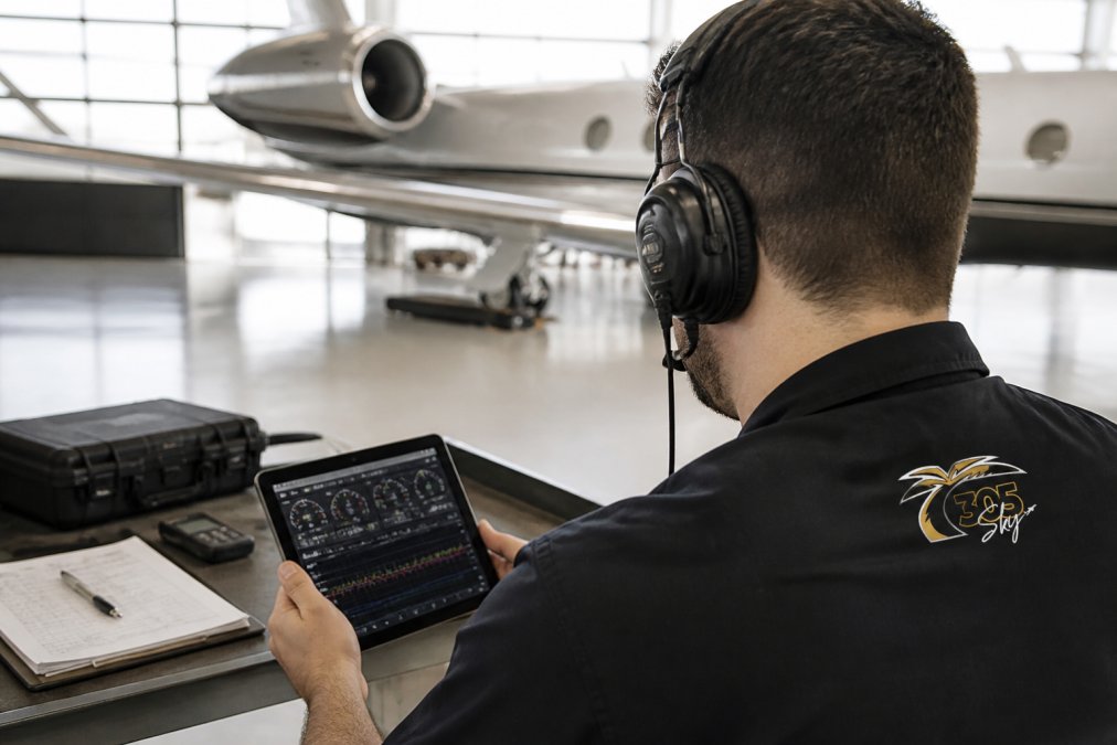 Aircraft maintenance technician monitoring real-time engine parameters on a digital display system in a modern hangar, wearing headset and reviewing diagnostic data on tablet with aircraft visible in background