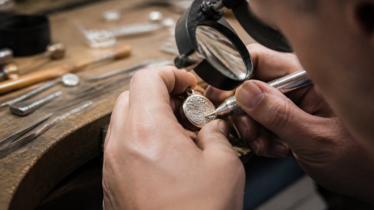 Jeweler's hands carefully working on a custom pendant with engraving tools and precision instruments in a well-lit workshop, magnifying glass showing detailed craftsmanship