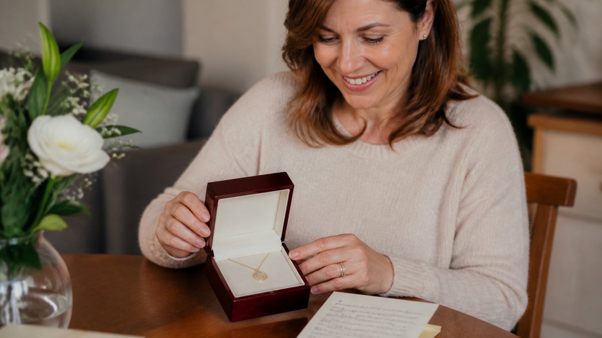 A woman in her 40s smiling as she opens a small mahogany-style gift box containing an engraved gold necklace, with a handwritten note beside it on a wooden table