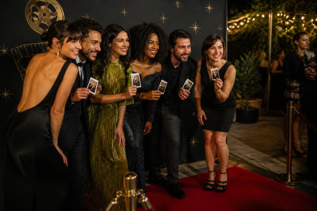 Guests in cocktail attire smiling and posing in an open-air photo booth with a branded step-and-repeat backdrop at an elegant film premiere after-party, instant prints in hand Guests in cocktail attire smiling and posing in an open-air photo booth with a branded step-and-repeat backdrop at an elegant film premiere after-party, instant prints in hand