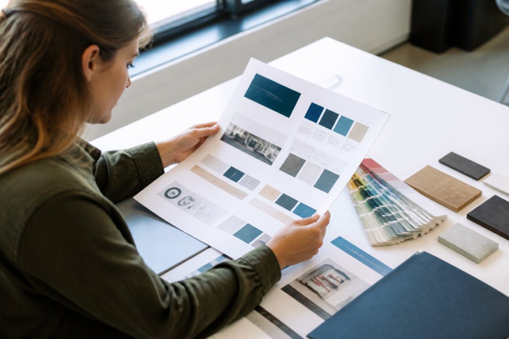 Professional designer reviewing printed brand guidelines document with color swatches, font samples, logo placement diagrams, and material samples spread across desk in bright office setting