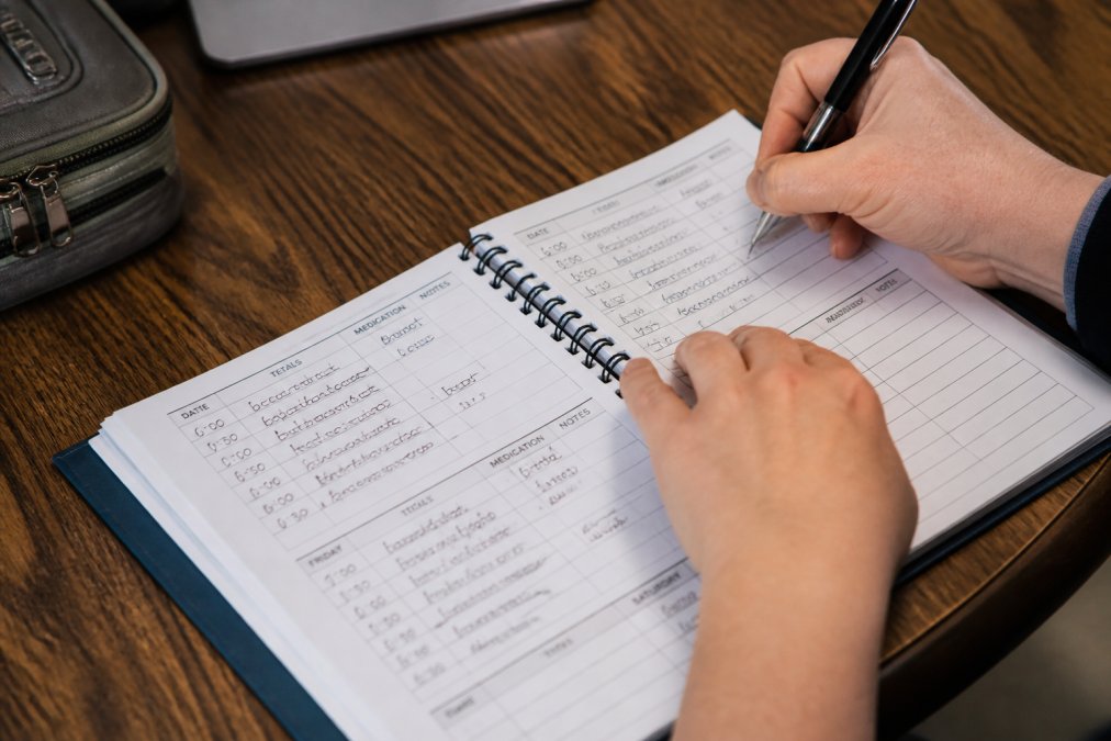 Close-up of hands writing in a care log notebook with a pen, showing organized notes and daily entries with clear handwriting and structured daily sections