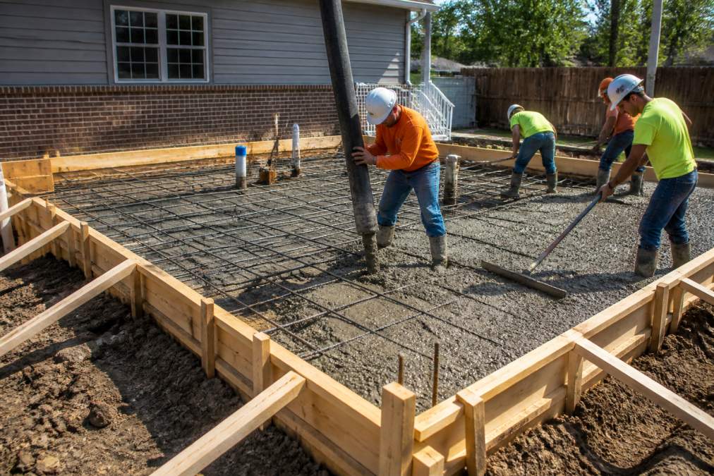 A construction crew in hard hats pouring concrete for a new home addition foundation, with a grid of rebar visible and wooden forms in place under bright morning sunlight