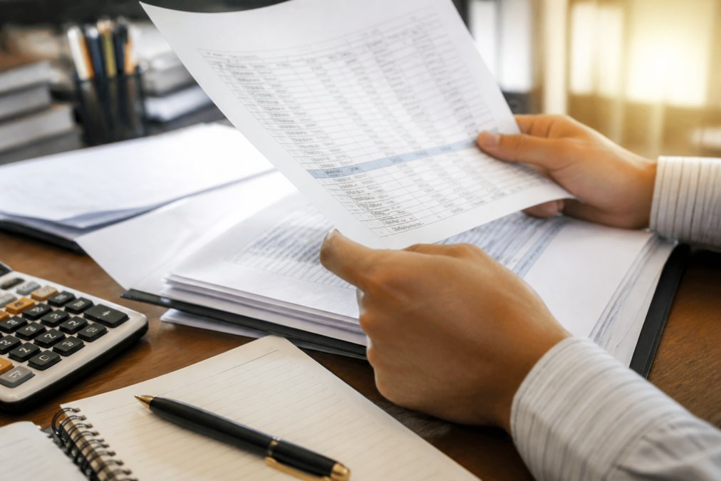 Close-up of hands holding financial statements and royalty documents, with a calculator and notepad visible on a workspace, under warm office lighting