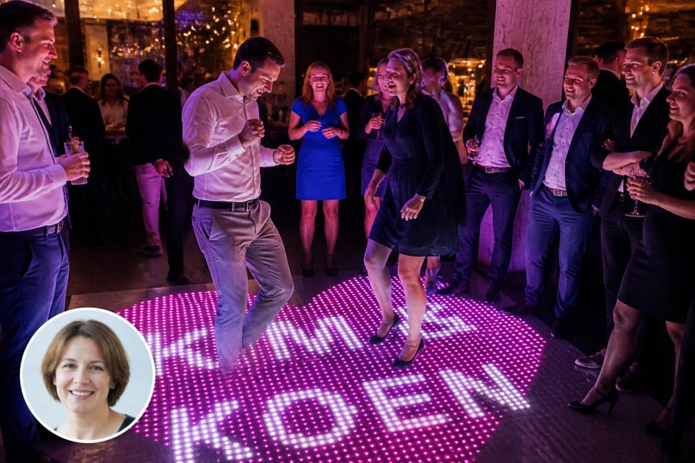 Groep diverse collega's die energiek dansen op een verlichte dansvloer met dynamische kleurveranderingen, terwijl anderen toekijken en glimlachen in een gezellige bedrijfsfeest-setting