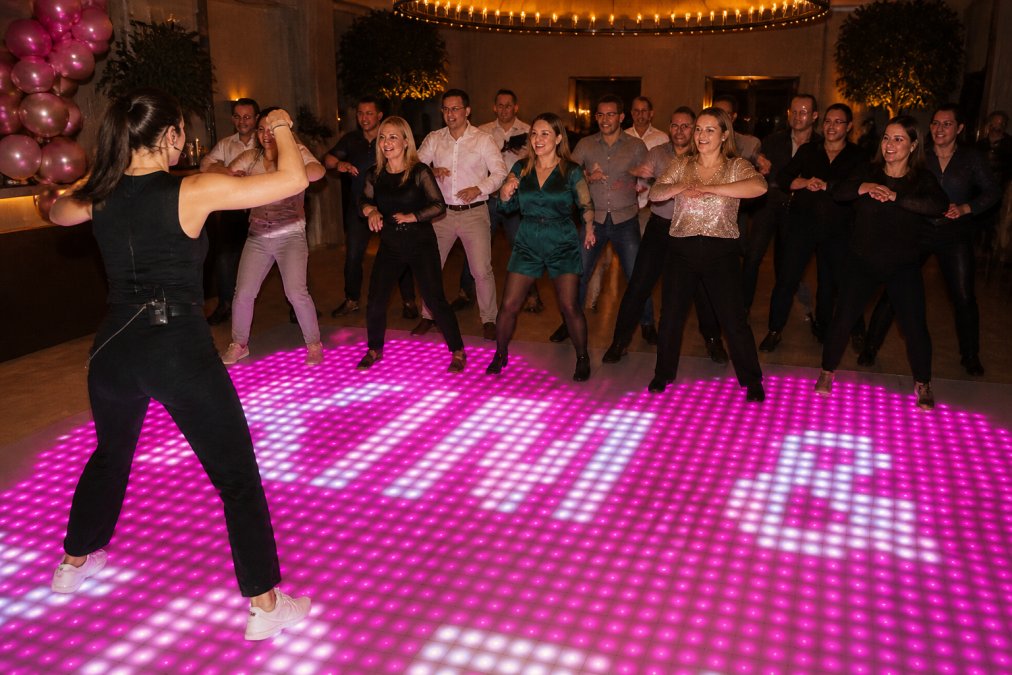 Professionele dansleraar die collega's begeleidt in een groepsdansles op een bedrijfsfeest, met vrolijke gezichten en beweging in een goed verlichte zaal