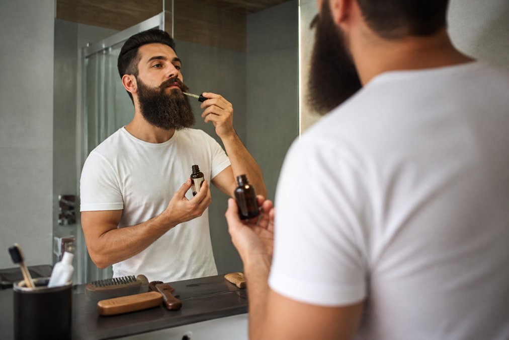 Un homme dans une salle de bain moderne appliquant de l'huile à barbe sur une barbe longue et fournie, avec un peigne en bois et un flacon d'huile visibles sur le comptoir