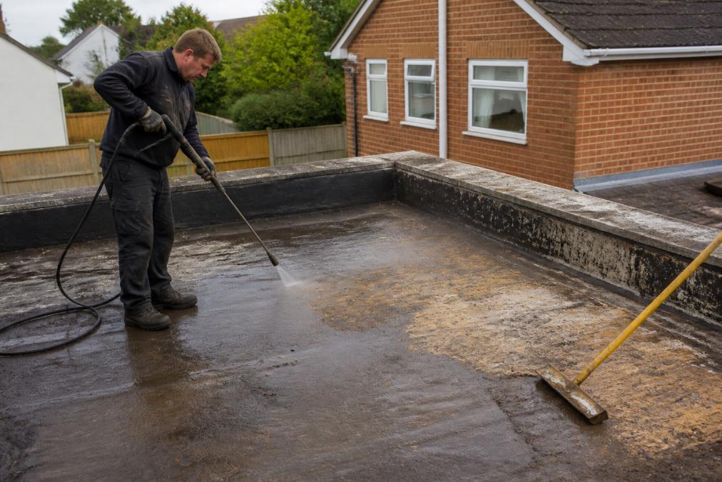 Professional roofer cleaning and preparing a flat roof deck with pressure washing equipment and removal tools, showing substrate exposed to bare surface in preparation for EPDM installation