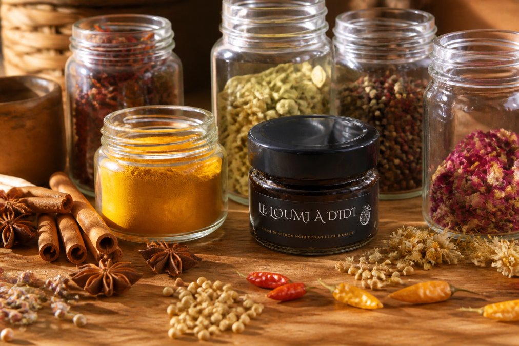 Assortiment coloré d'épices artisanales en pots de verre et bocaux, disposés sur une table en bois avec des graines, fleurs et bâtons d'épices rouges et jaunes visibles, lumière naturelle chaude dorée