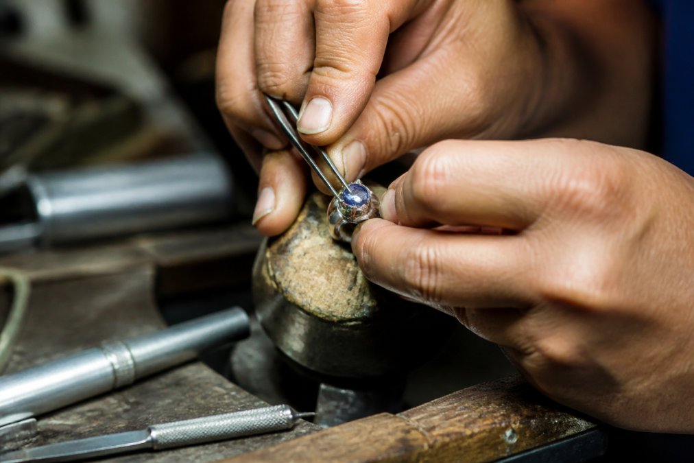 Close-up of a jeweler's hands using specialized tools to set a gemstone into a sterling silver band, with fine detail work visible under bright workshop lighting