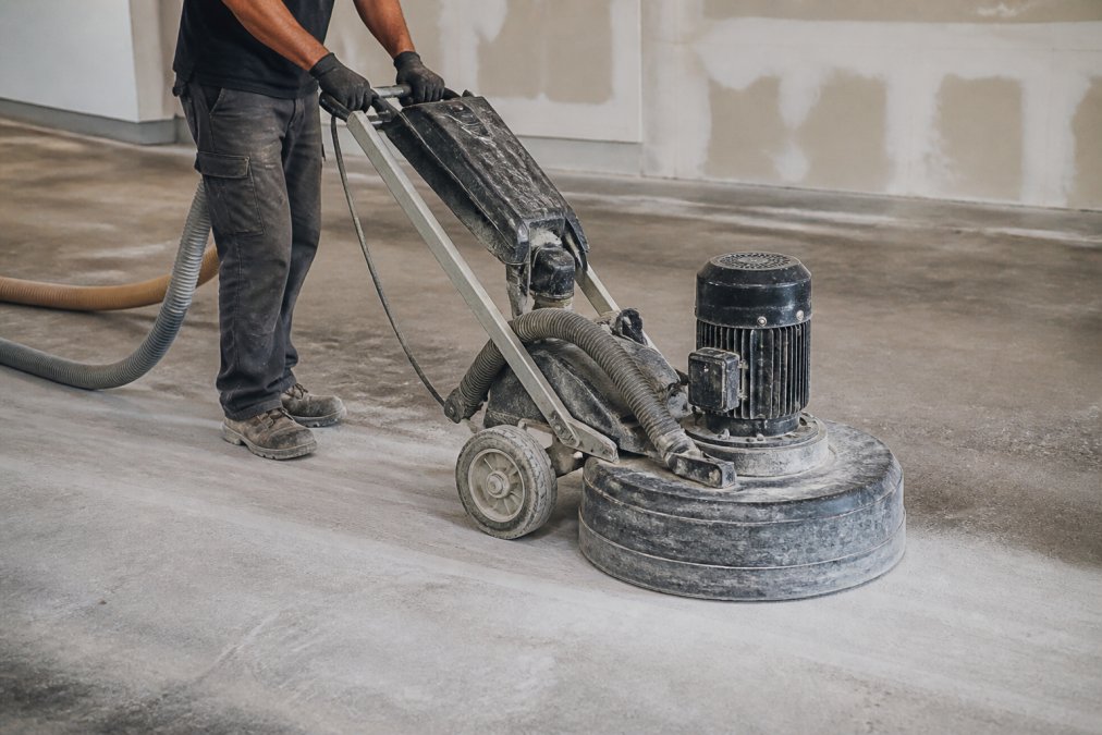 Professional concrete grinder with diamond tooling in action on concrete floor surface, showing dust collection system and proper operator stance with safety equipment