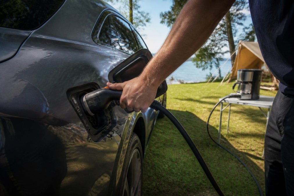 Close-up of a hand connecting a V2L adapter to an electric car's charging port outdoors, with the cable running to a camping appliance on a sunny day