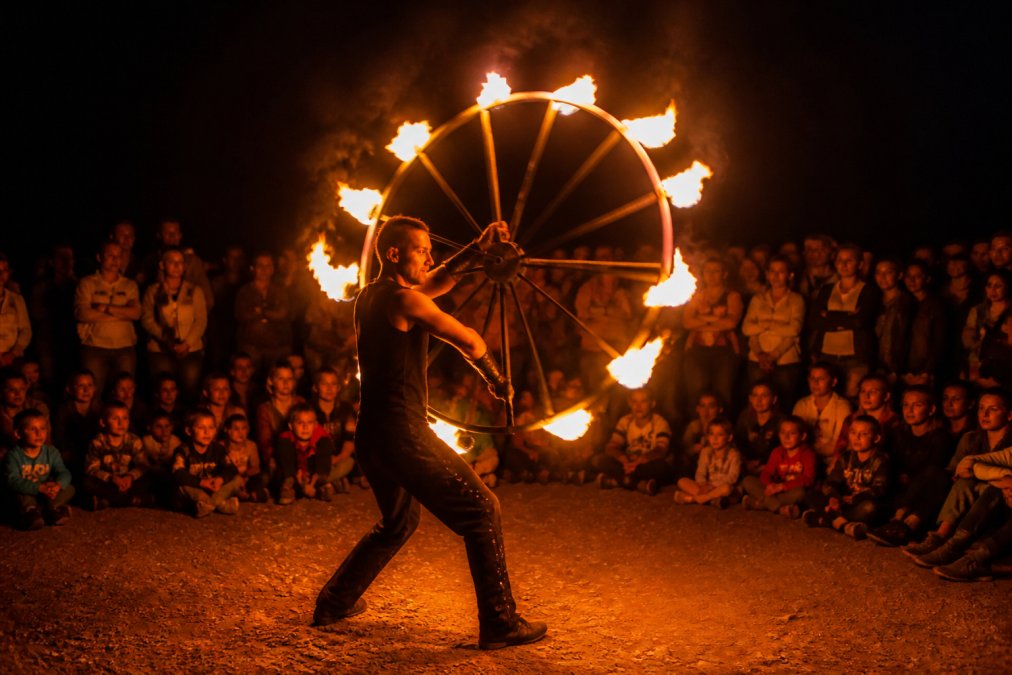 Erfaren ildkunstner udfører spektakulært ildshow ved aftenstid med flammende ildhjul, publikum samlet i mørket omkring kunstneren, varmt lys fra ilden belyser ansigterne