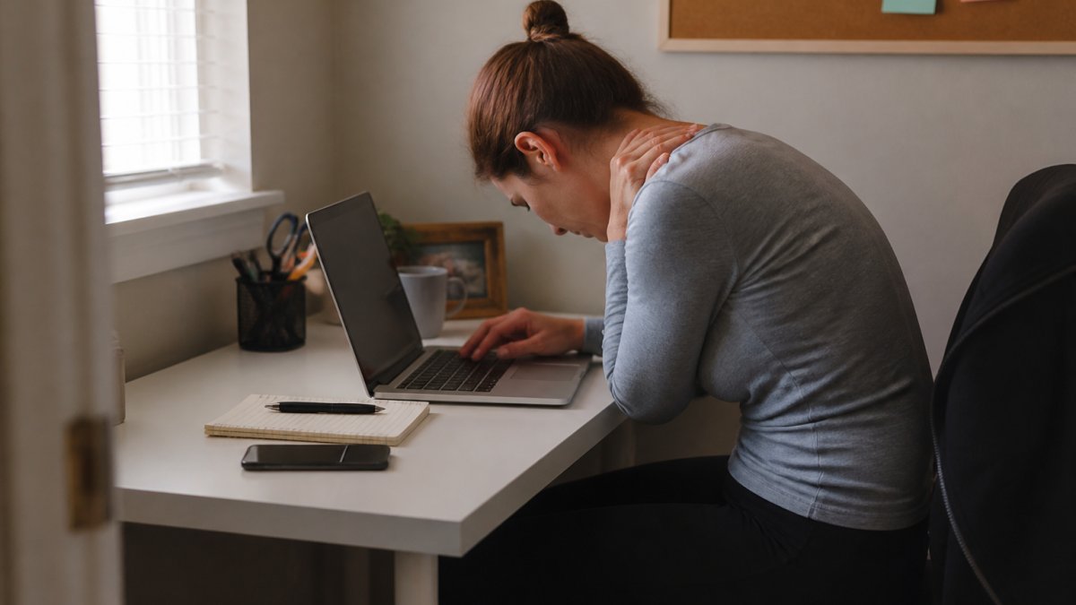 Remote worker hunched over laptop at cramped desk, showing poor posture and visible neck and shoulder strain in a small home office