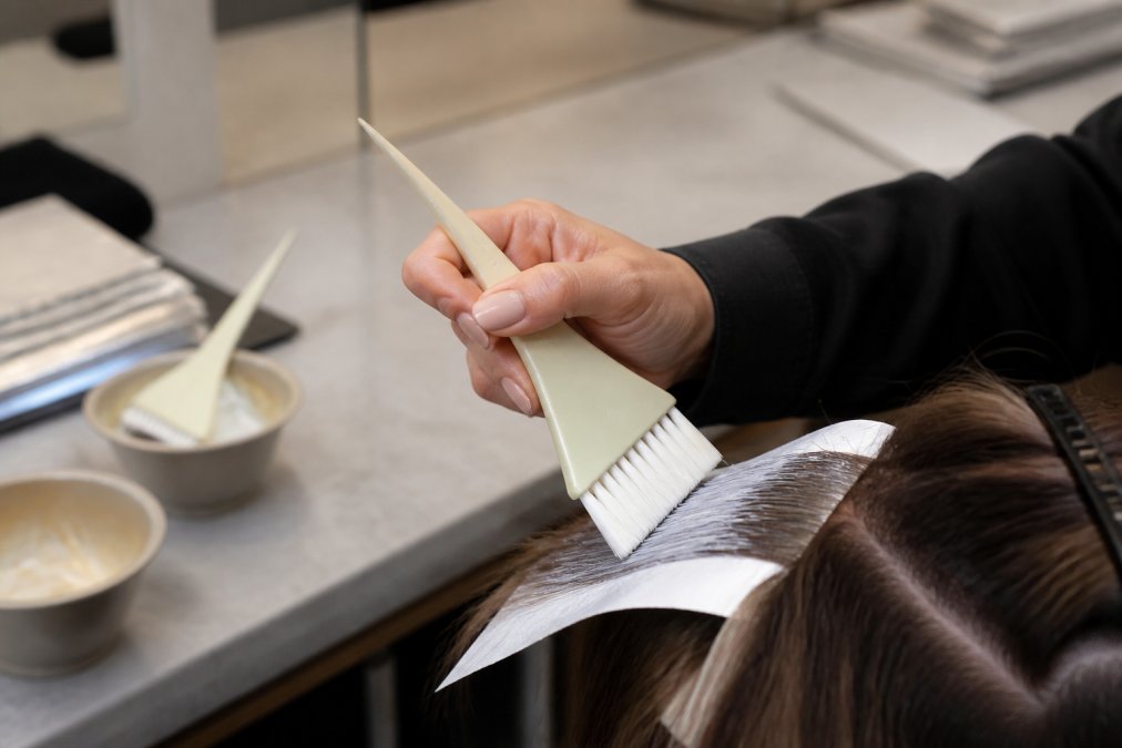 Professional colorist's hand holding an ergonomic feathered brush with proper thumb-grip positioning during a tint application on sectioned hair, demonstrating correct finger placement and wrist alignment