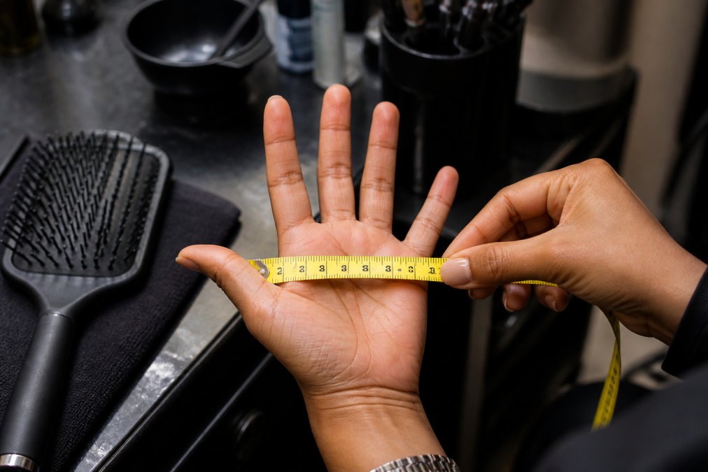 Close-up of a person's hand holding a measuring tape across their palm width, with a hair brush resting on a salon workstation nearby