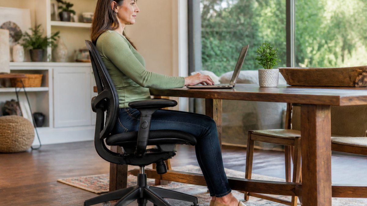 Person sitting in an ergonomic office chair with proper lumbar support, feet flat on the floor, demonstrating correct neutral sitting posture at a desk with natural office lighting