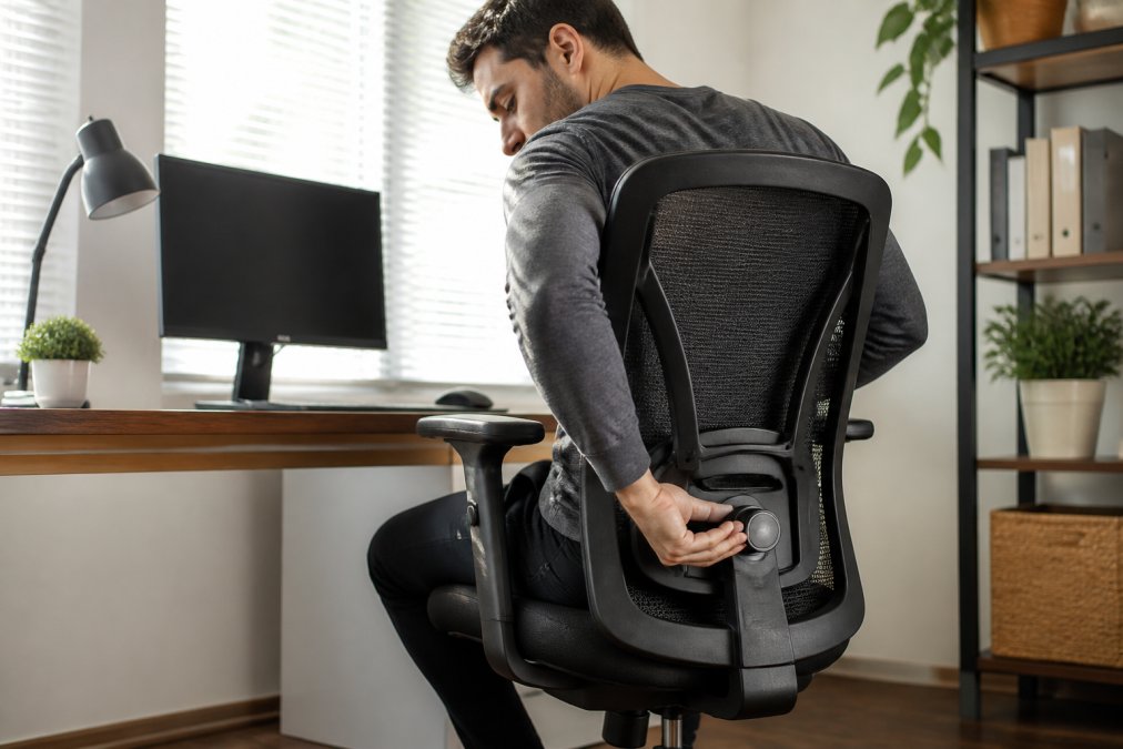 A person adjusting the lumbar support knob on an ergonomic office chair in a bright home office, with a desk and monitor visible in the background