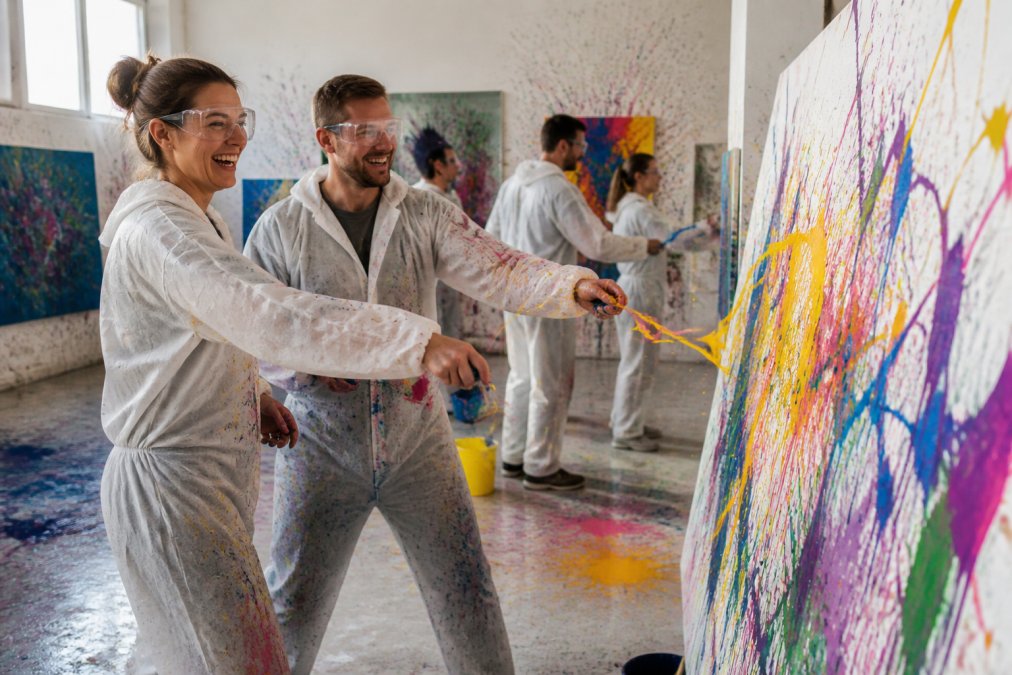 Ein Paar wirft lachend Farbe auf eine Leinwand bei einem kreativen Workshop als Erlebnisgeschenk für Paare.
