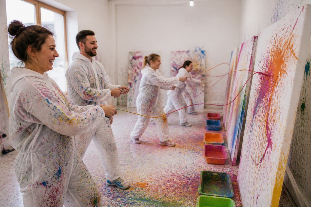 Eine Gruppe von Menschen lacht und wirft bunte Farbe auf große Leinwände in einem hellen Workshop-Studio, alle tragen farbbesprenkelte Schutzkleidung
