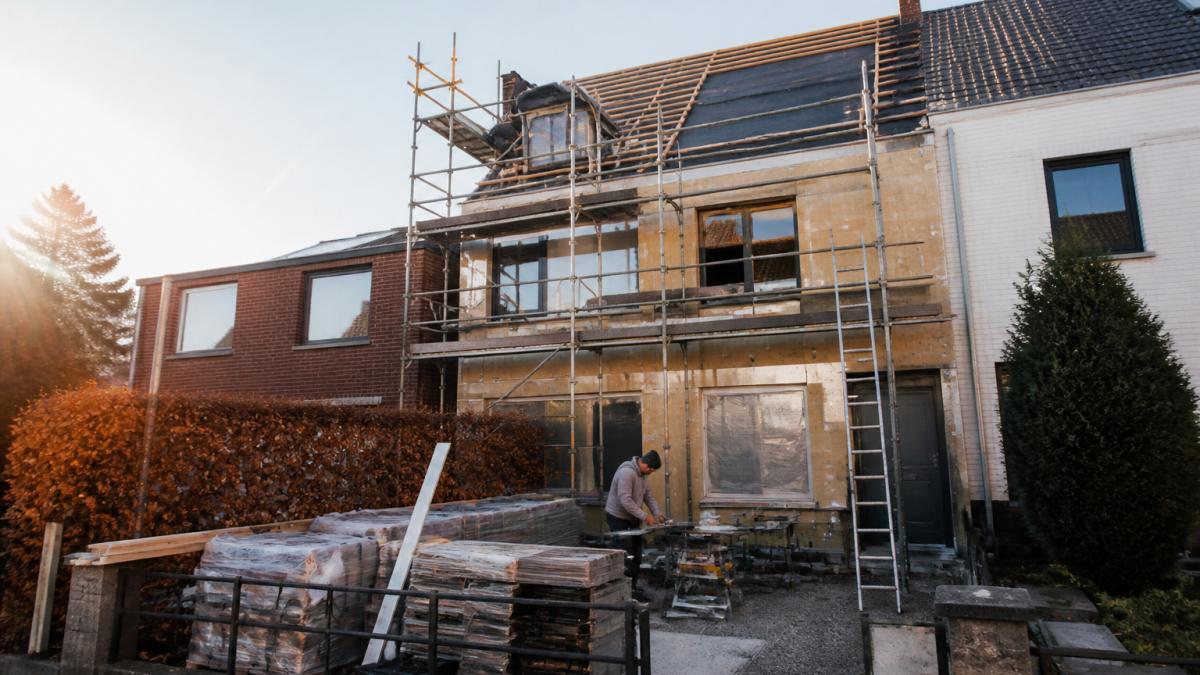 Een renovatiewerf in een rijwoning waar een ploeg vakmensen dak- en gevelwerken uitvoert, met steigers en materiaal in de voortuin, ochtendlicht