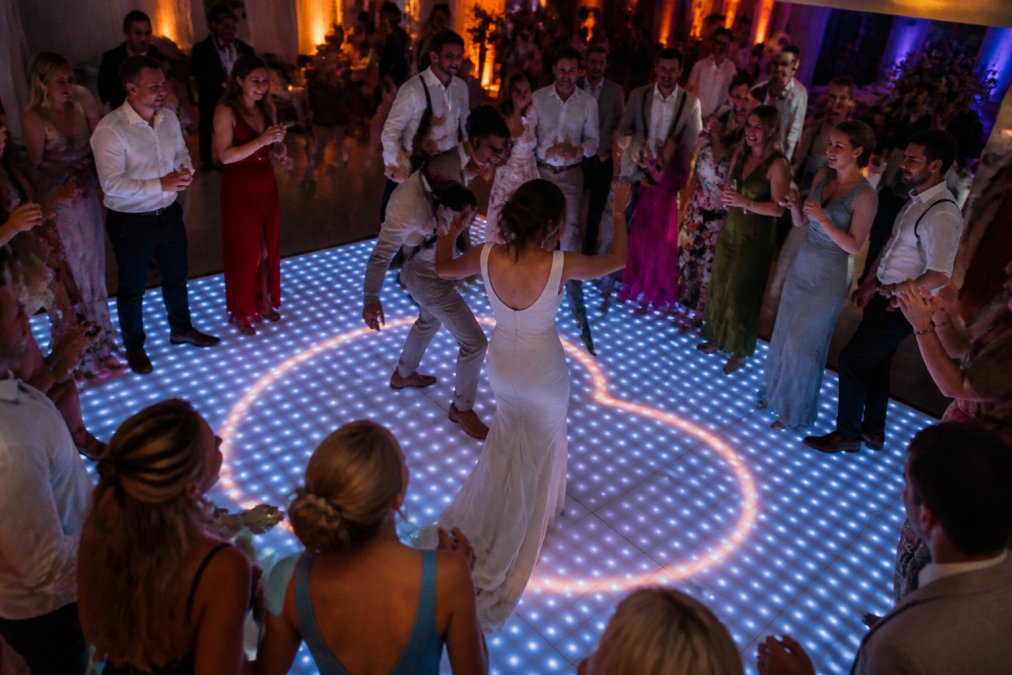 Een verlichte dansvloer met een hart en namen erin geprogrammeerd, gasten dansen eromheen op een bruiloftsfeest, warme sfeerverlichting op de achtergrond