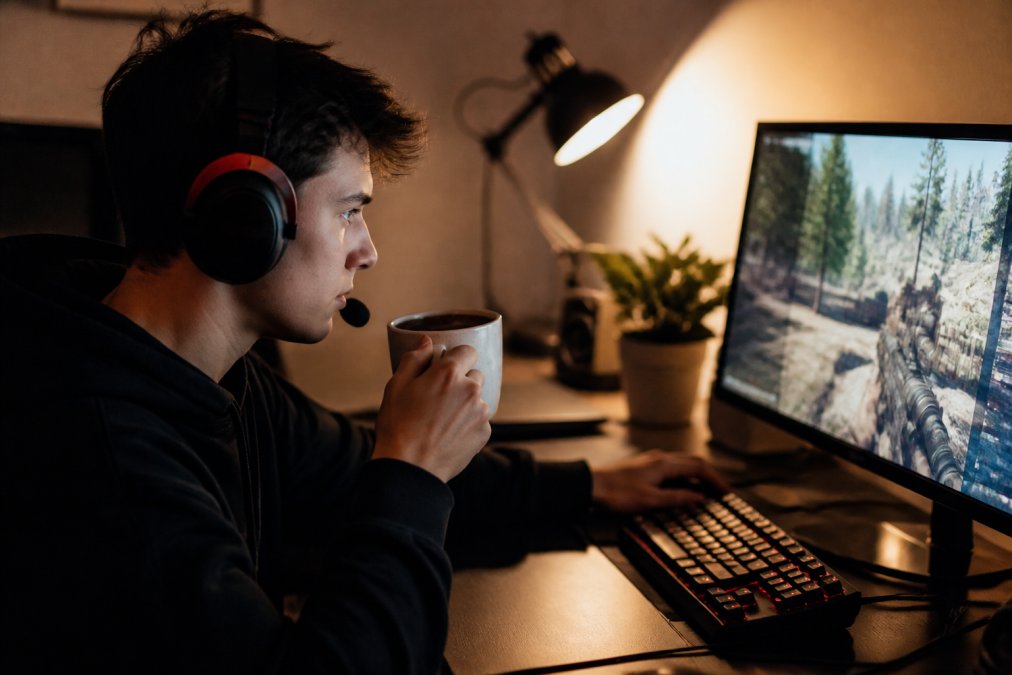 A young gamer at a desk with a headset, holding a ceramic mug of coffee, looking focused at a monitor with a game on screen, warm desk lamp lighting