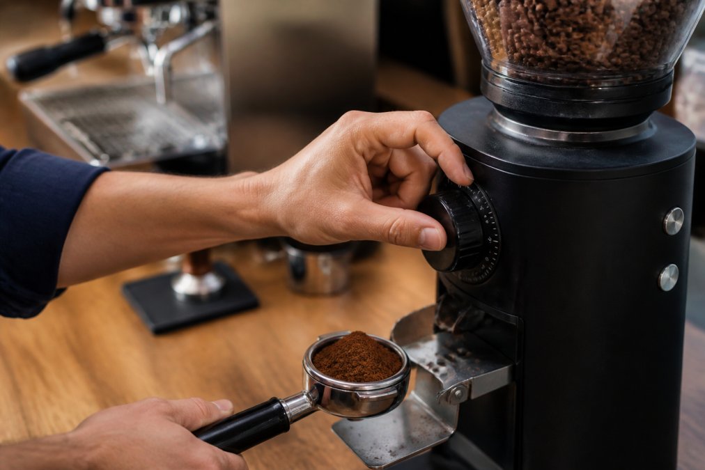 Close-up of a home barista's hands adjusting the dial on a flat burr coffee grinder, with a portafilter holding freshly ground coffee nearby on a counter, soft natural light