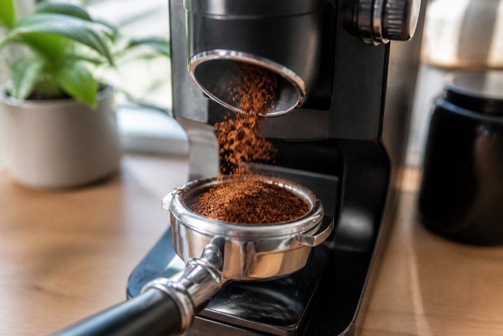 Close-up of freshly ground espresso coffee falling into a portafilter basket, with a modern electric grinder in the background and soft natural light on a kitchen counter