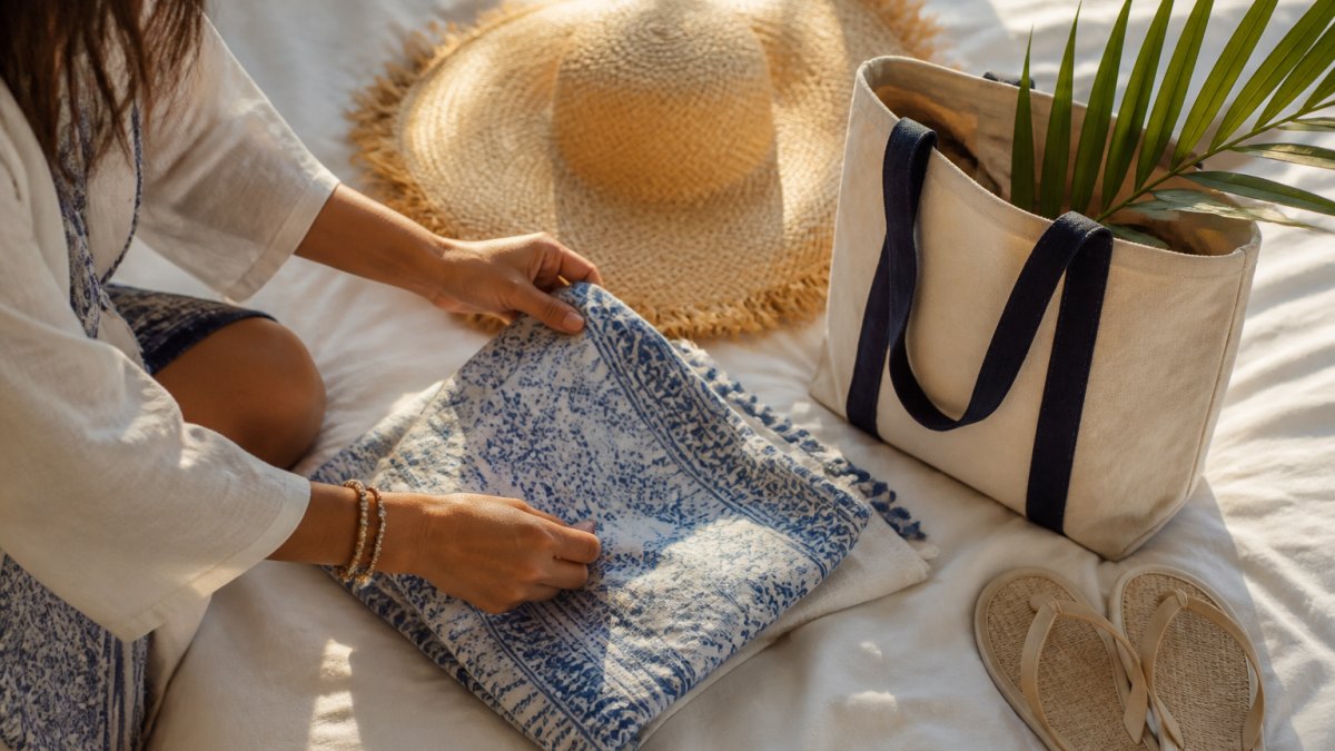 Wide-brim straw hat, cotton sarong, and canvas tote bag arranged on white linen with a tropical greenery sprig.