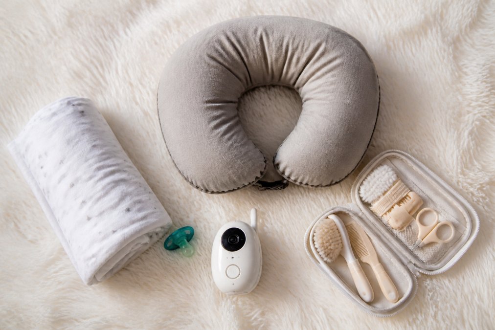 A neatly organized nursery flat lay showing a swaddle blanket, nursing pillow, pacifier, baby monitor, and small grooming kit arranged on a soft white surface, photographed from above with warm natural window light