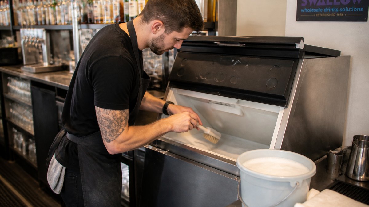 A bar worker in a clean apron using a soft brush to clean the interior of a commercial ice machine, with a bucket of warm soapy water and clean cloths nearby in a well-lit back bar area