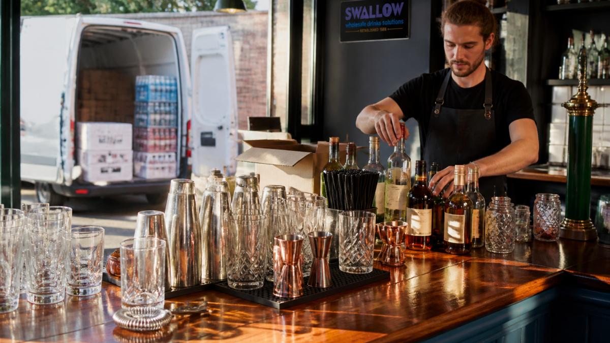 Professional bartender arranging bottles, glassware, and bar tools on a polished wooden counter with a white van delivery box visible in the background, natural afternoon light streaming through the venue