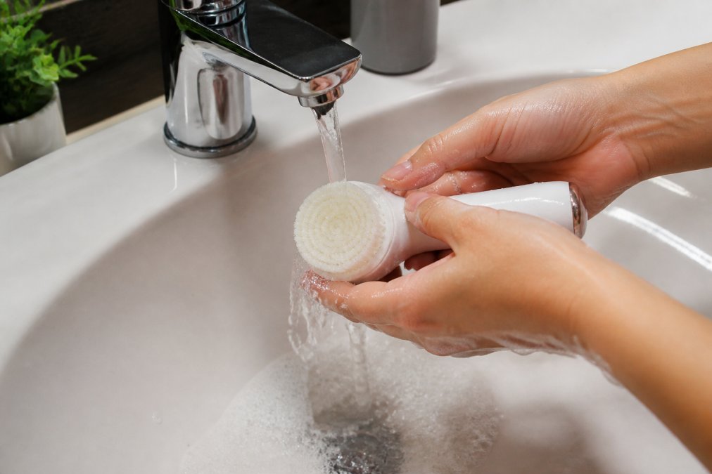 Hands holding facial cleansing brush under running water with soft brush bristles visible, demonstrating gentle cleaning technique over white bathroom sink with soap suds