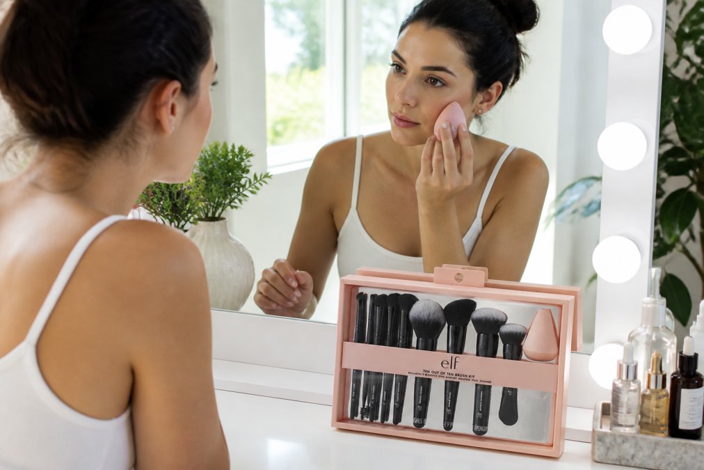 Woman applying foundation to face with ergonomic-handled beauty sponge at well-lit vanity mirror, demonstrating proper stippling technique with natural window lighting