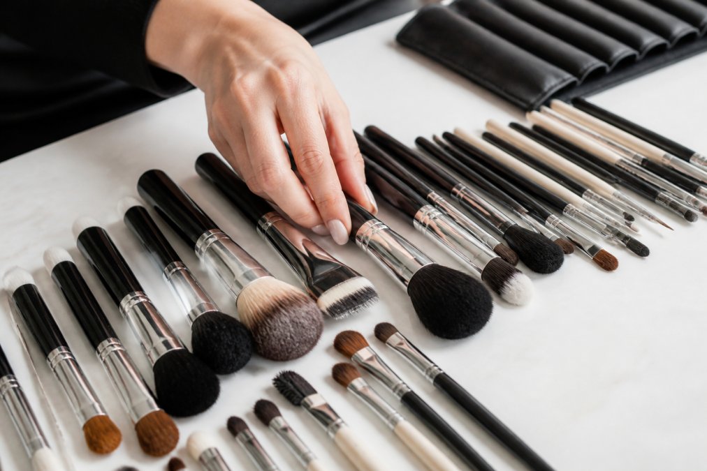 Professional makeup artist's hand arranging an array of essential brushes on a clean white surface, showing different brush shapes and sizes organised by type with natural studio lighting