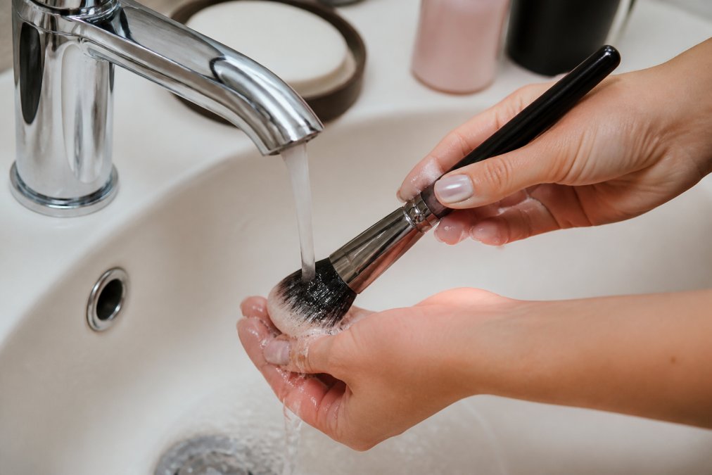 Close-up of hands rinsing makeup brushes under running water with a gentle cleanser, showing proper technique and care with warm water flowing over the bristles