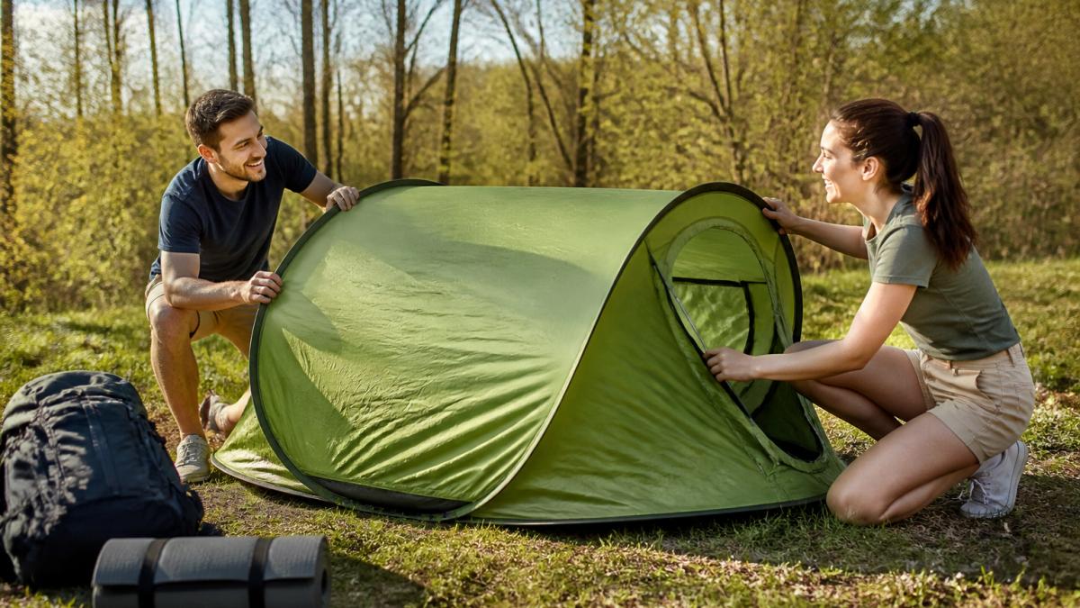 A young couple pitching a green pop-up tent at a sunny campsite, with a backpack and sleeping pad nearby, smiling as they set up