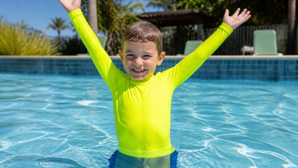 Young child wearing a properly fitted long-sleeve rash guard in a bright neon color, standing in shallow pool water with arms raised showing full range of motion and snug but comfortable coverage